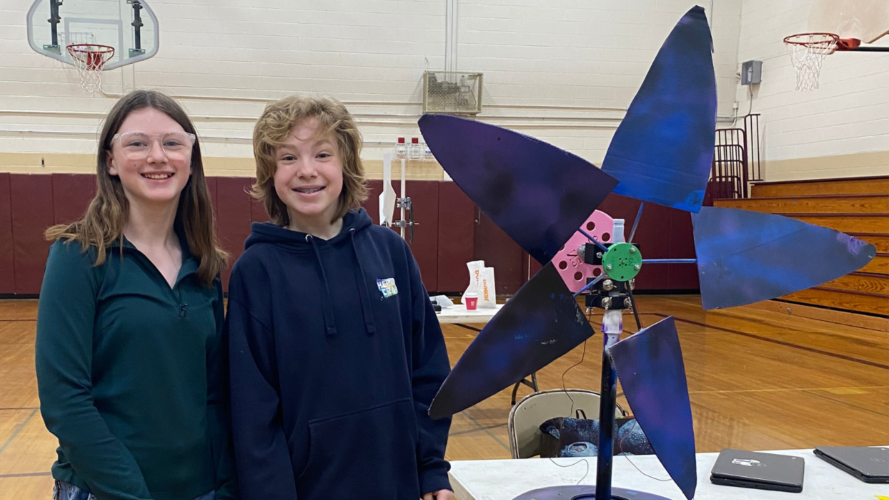 Two students stand beside a large windmill project painted purple and blue